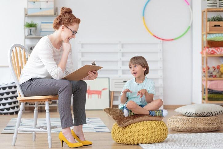 Cheerful young kid talking with helpful child counselor during psychotherapy session in children mental health center